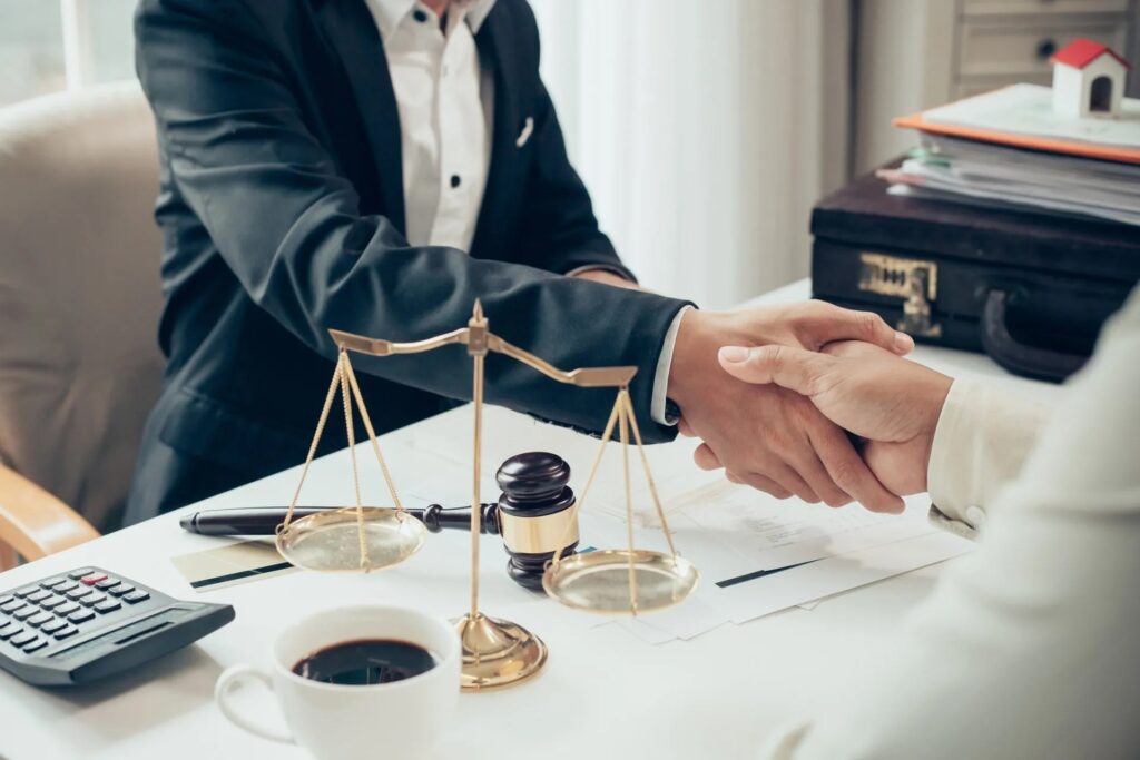 Two people handshaking over a desk with a gavel, a scale of justice and cup of coffee beside them