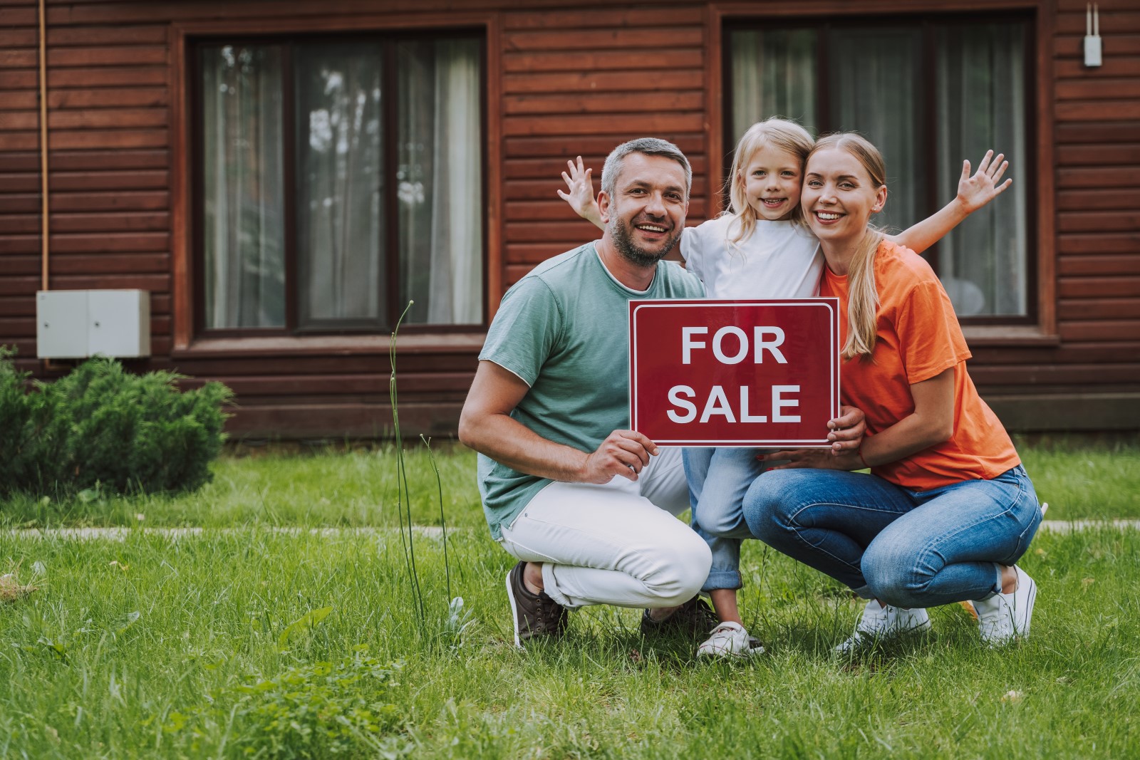 Family of three holding a red for sale sign in front of a brown wood paneled home