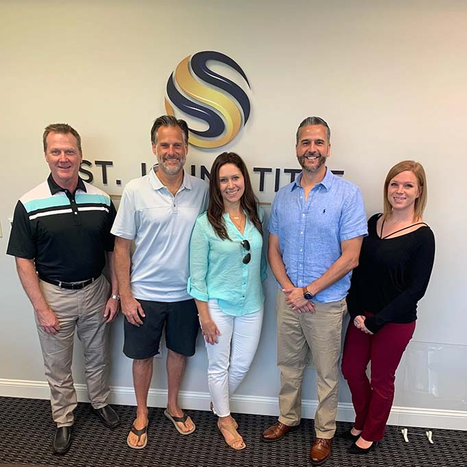 Five people standing and smiling in front of the St Johns Title logo in an office